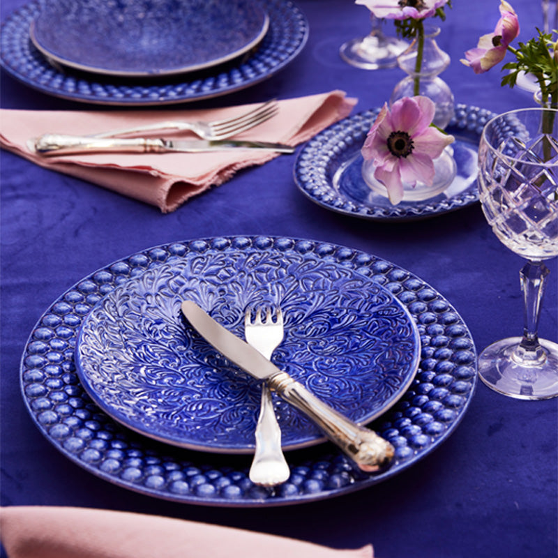 Blue ceramic plates with floral patterns on a purple tablecloth, accompanied by silver cutlery and pink napkins.