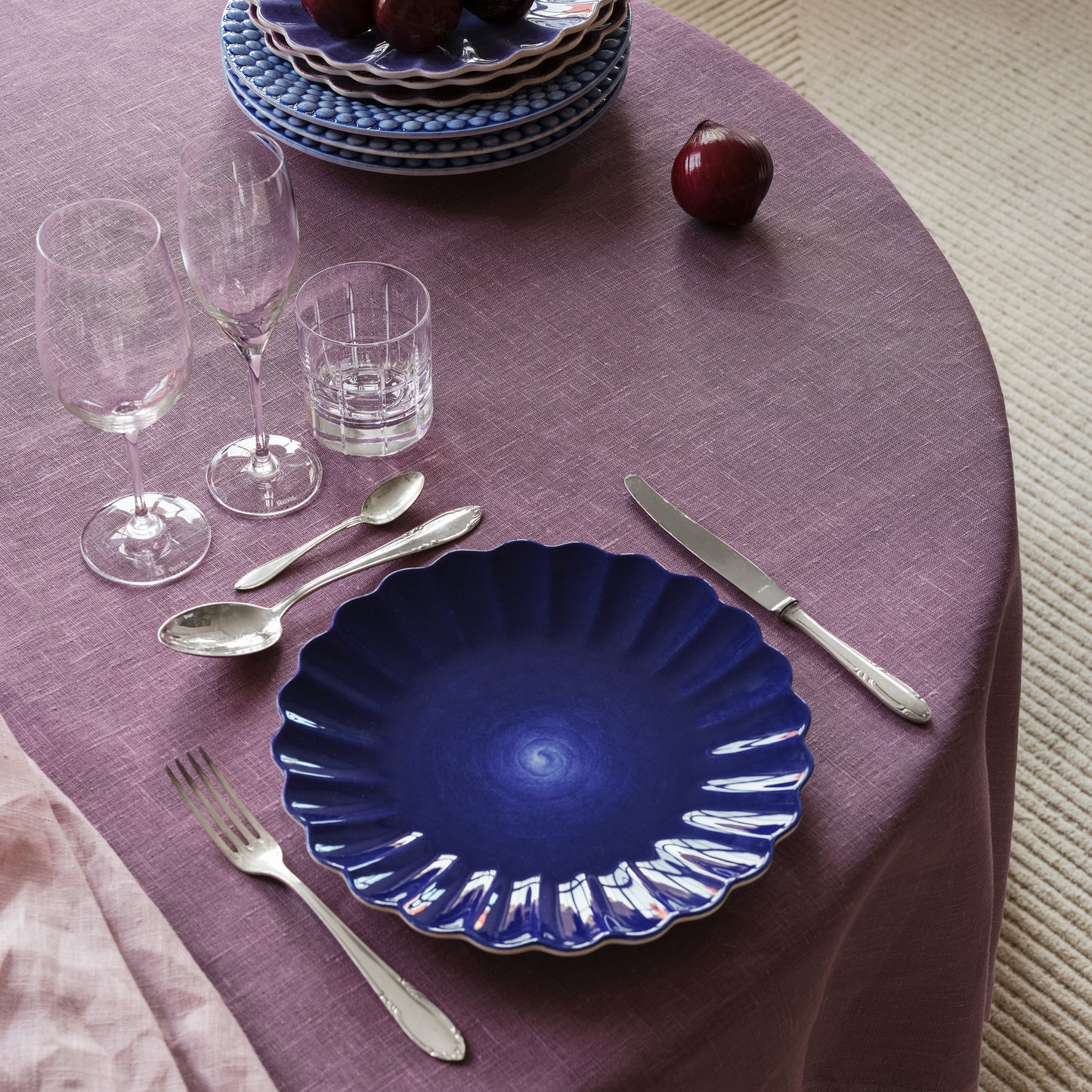 Dinner table setting with blue plates, silverware, and glasses on a purple tablecloth.
