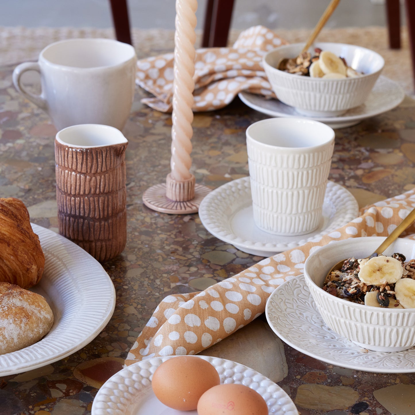 Breakfast table setting with cups, bowls, and pastries on a patterned tablecloth.
