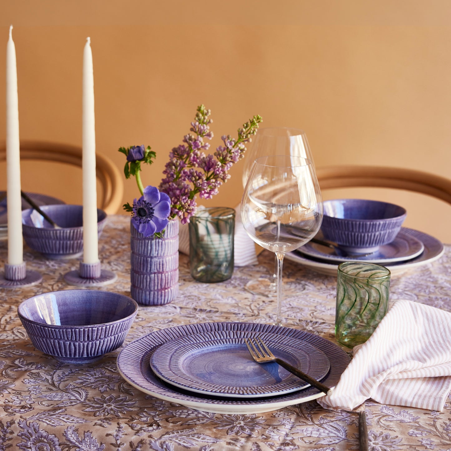 Elegant dining table setting with purple plates, bowls, and glasses on a patterned tablecloth.