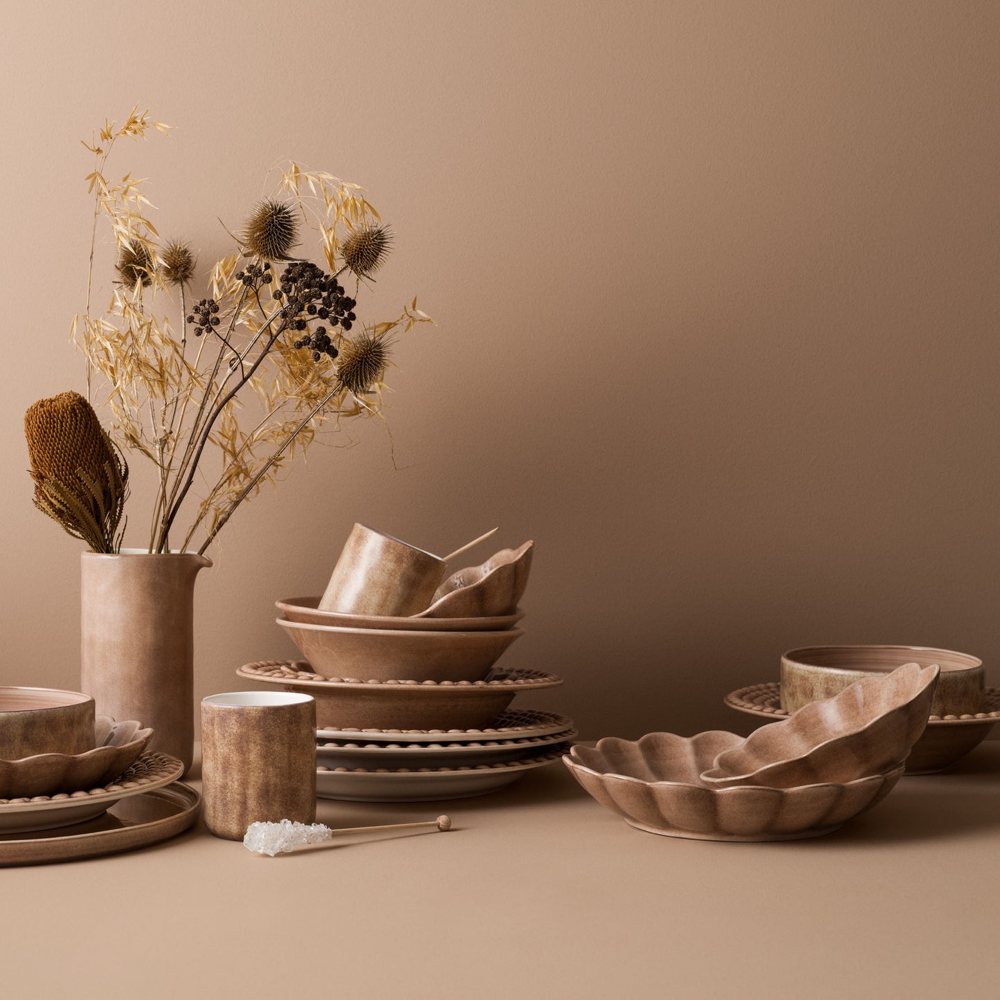 Set of brown ceramic dishes and a vase with dried plants on a beige surface.