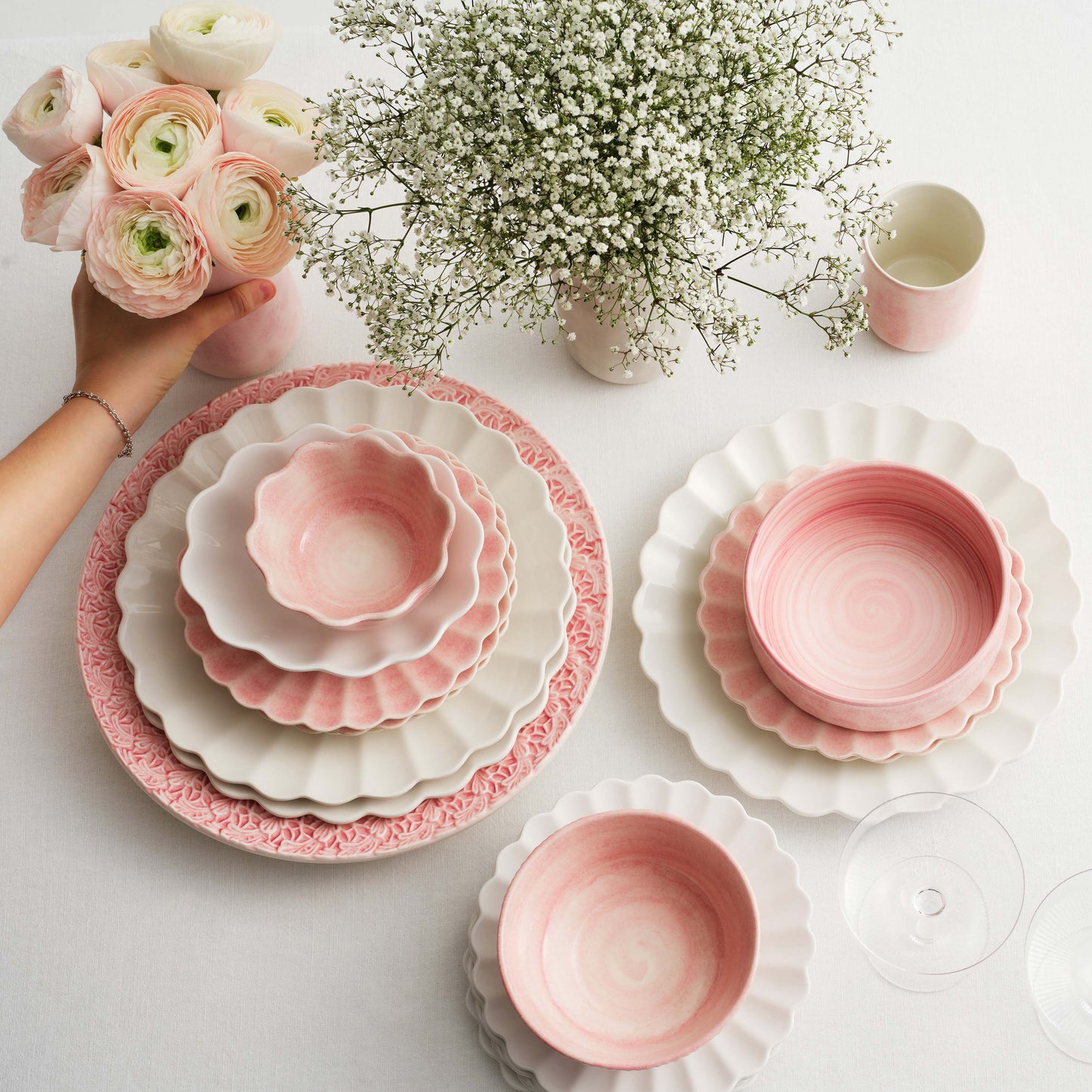Pink and white ceramic dishes on a table with flowers and a hand holding a bouquet.