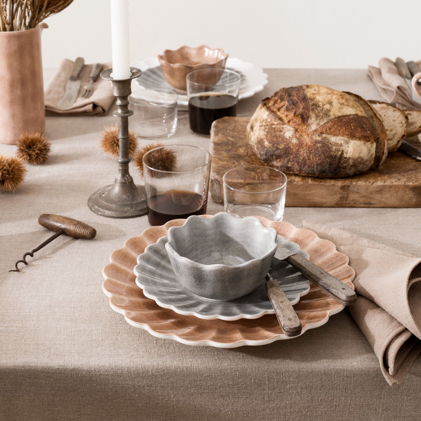 Dining table setting with ceramic bowls, glasses, bread, and candles on a neutral background