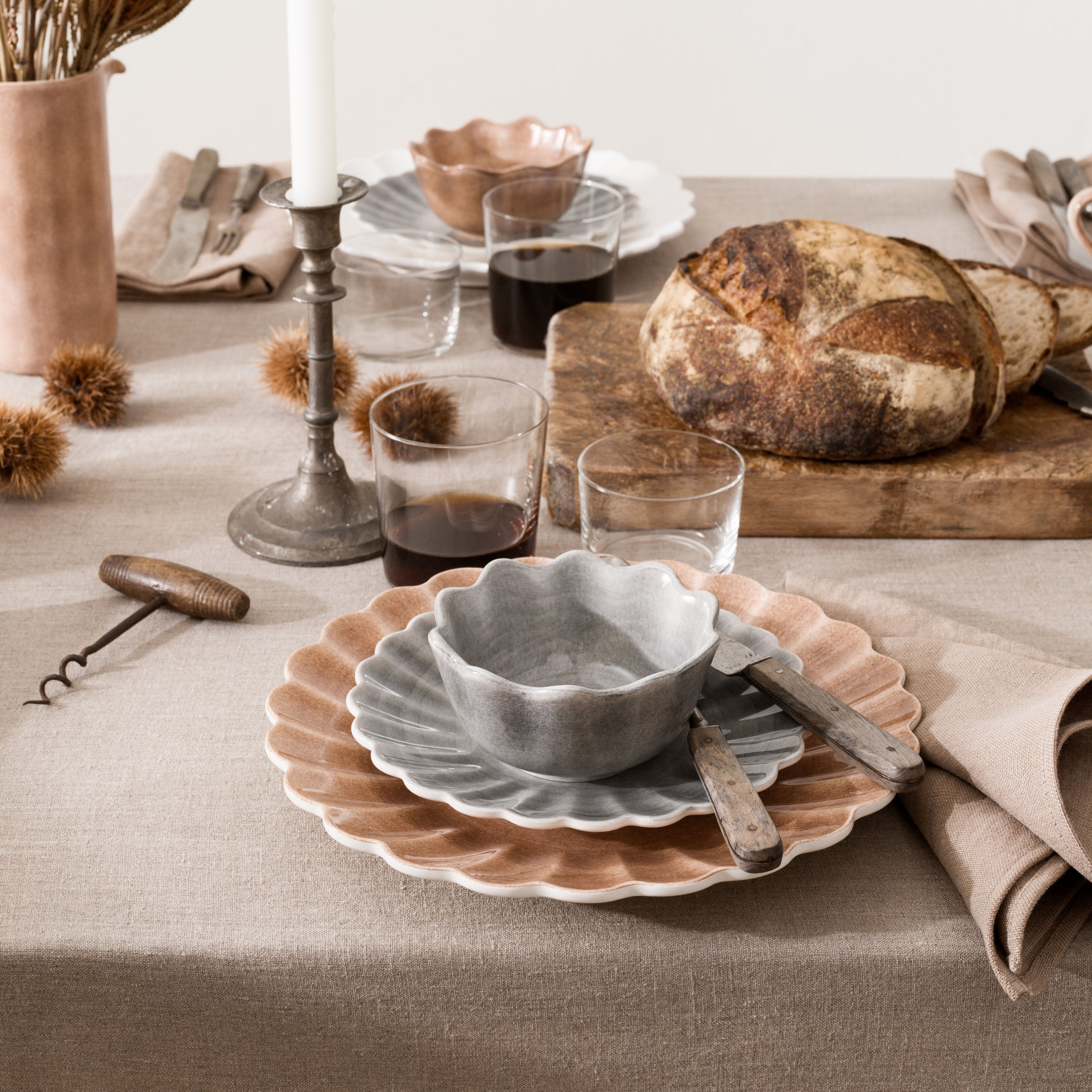 Dining table setting with ceramic bowls, glasses, bread, and candles on a neutral background