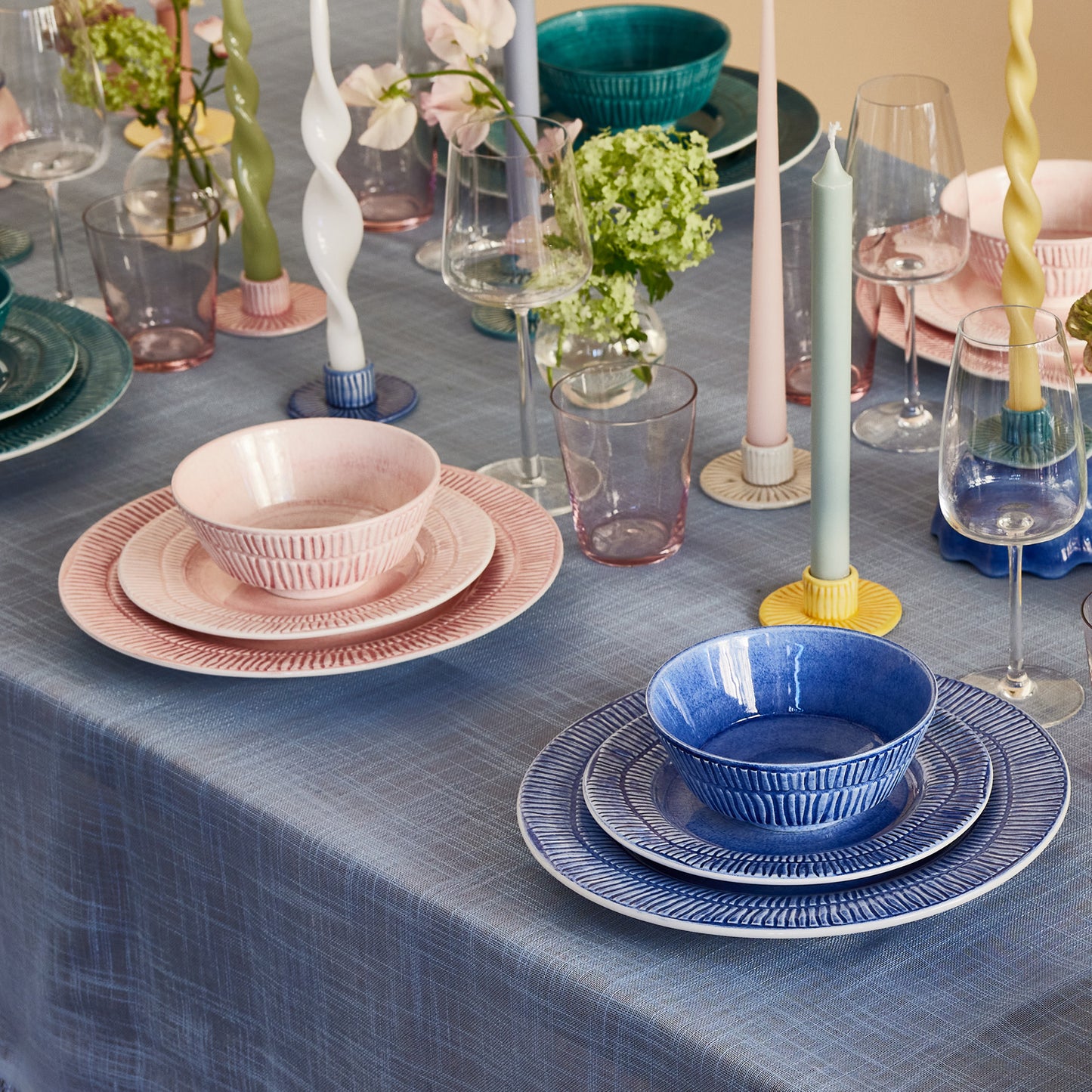 Dining table setting with blue and pink dishes, candles, and flowers on a blue tablecloth.