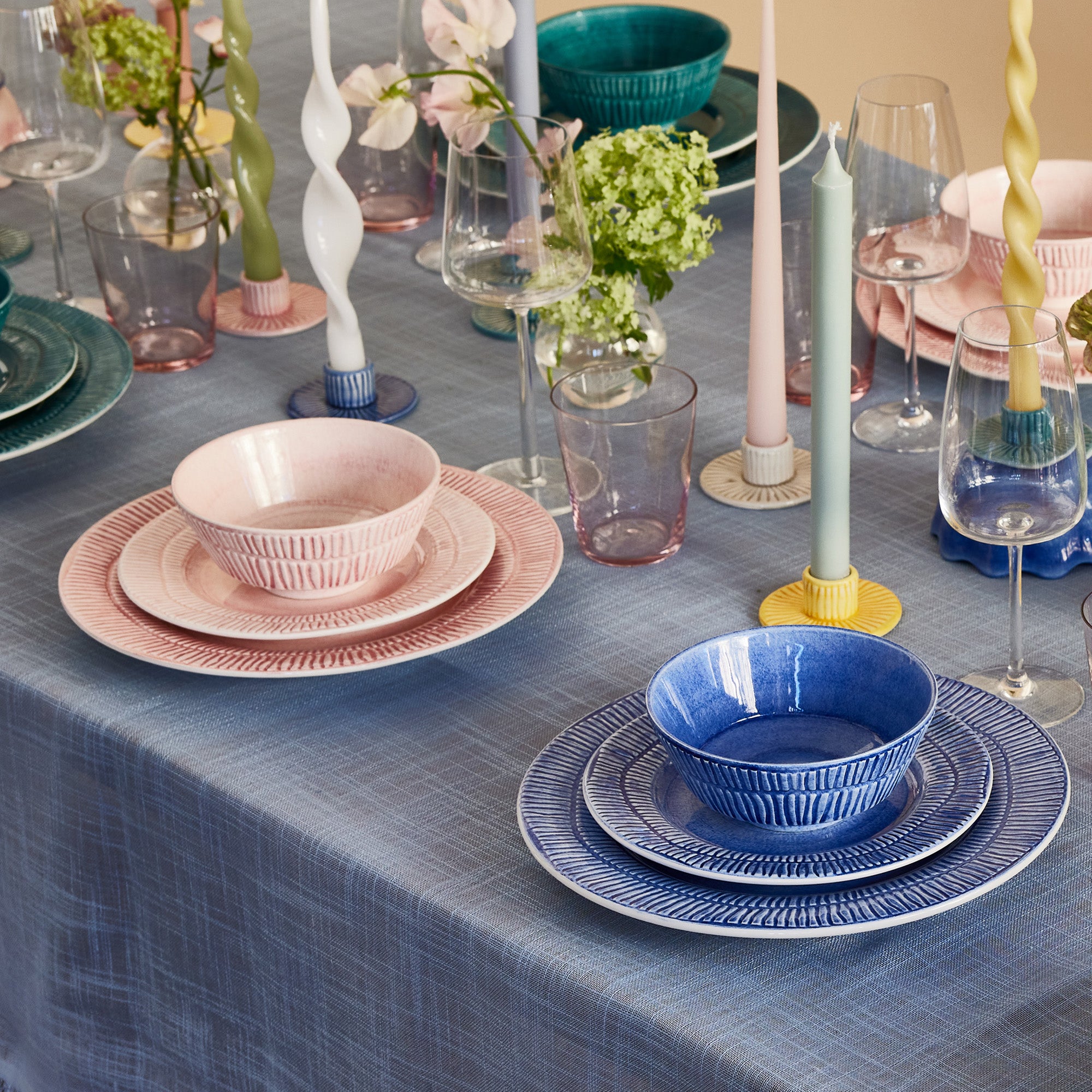 Dining table setting with blue and pink dishes, candles, and flowers on a blue tablecloth.