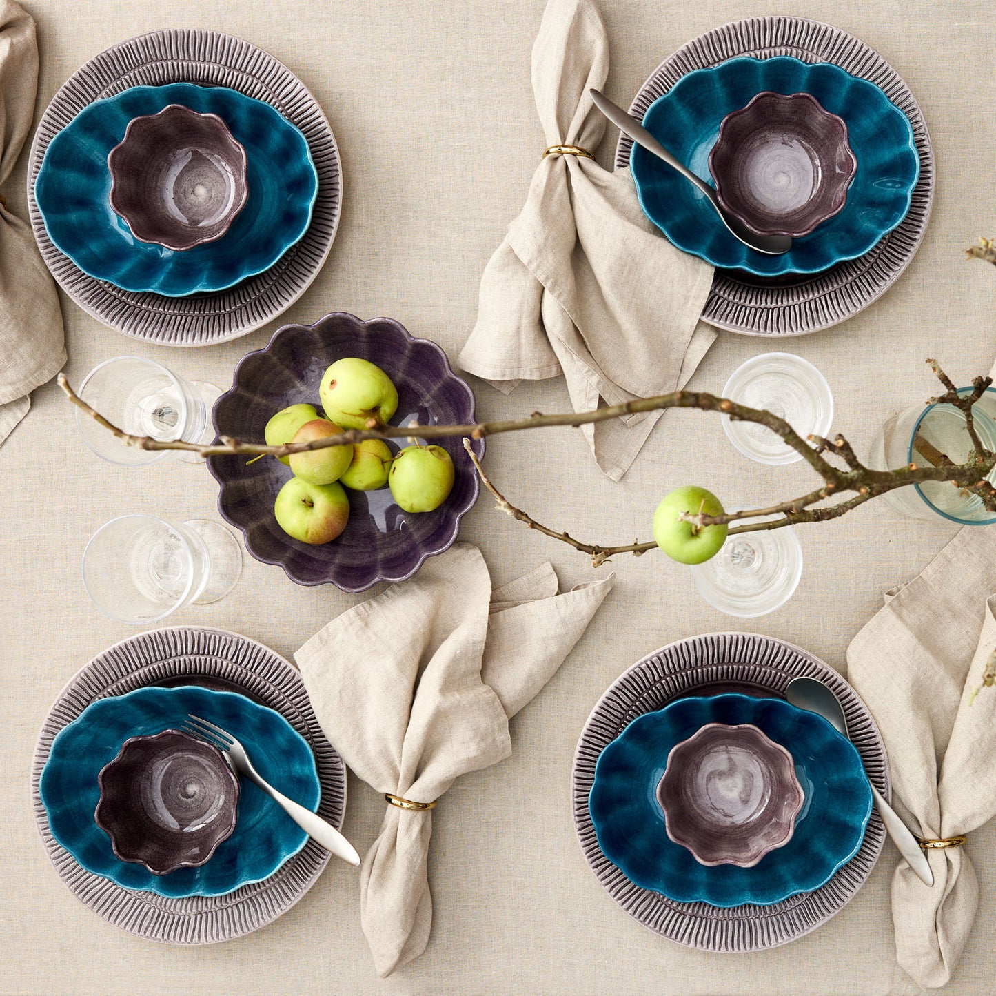 Table setting with blue and purple ceramic plates, green apples, and branches on a beige tablecloth.