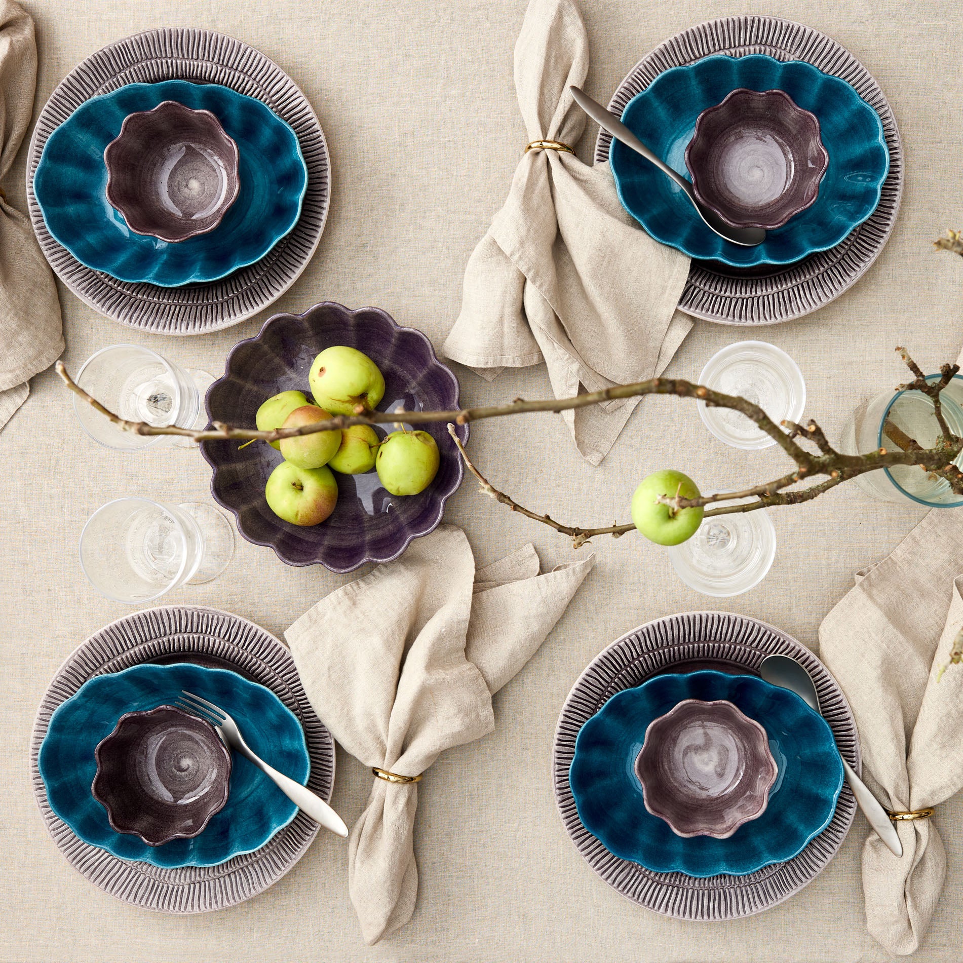Table setting with blue and purple ceramic plates, green apples, and branches on a beige tablecloth.
