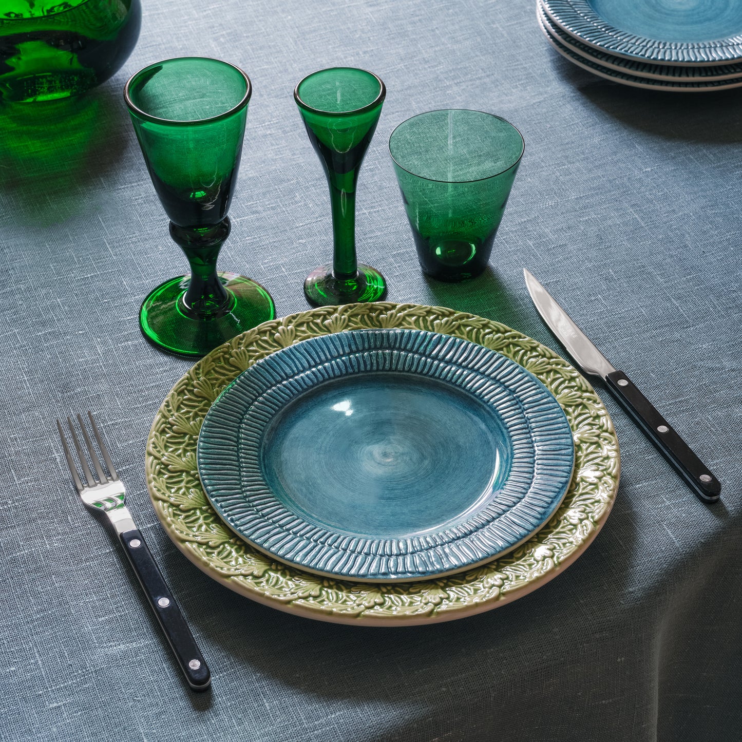 Table setting with green glasses, blue plate, and silverware on a gray tablecloth.
