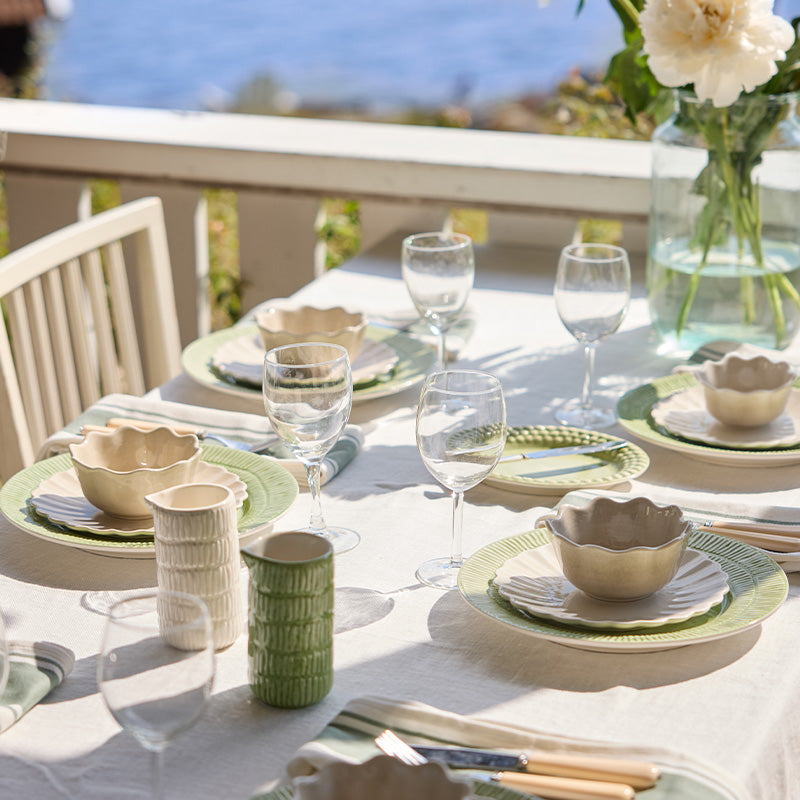 Dining table set with green and white dishes, glasses, and a vase with flowers on a balcony.