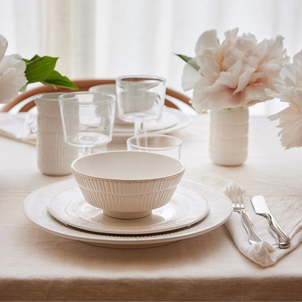 Dining table setting with white ceramic dishes, glasses, and silverware on a beige tablecloth.