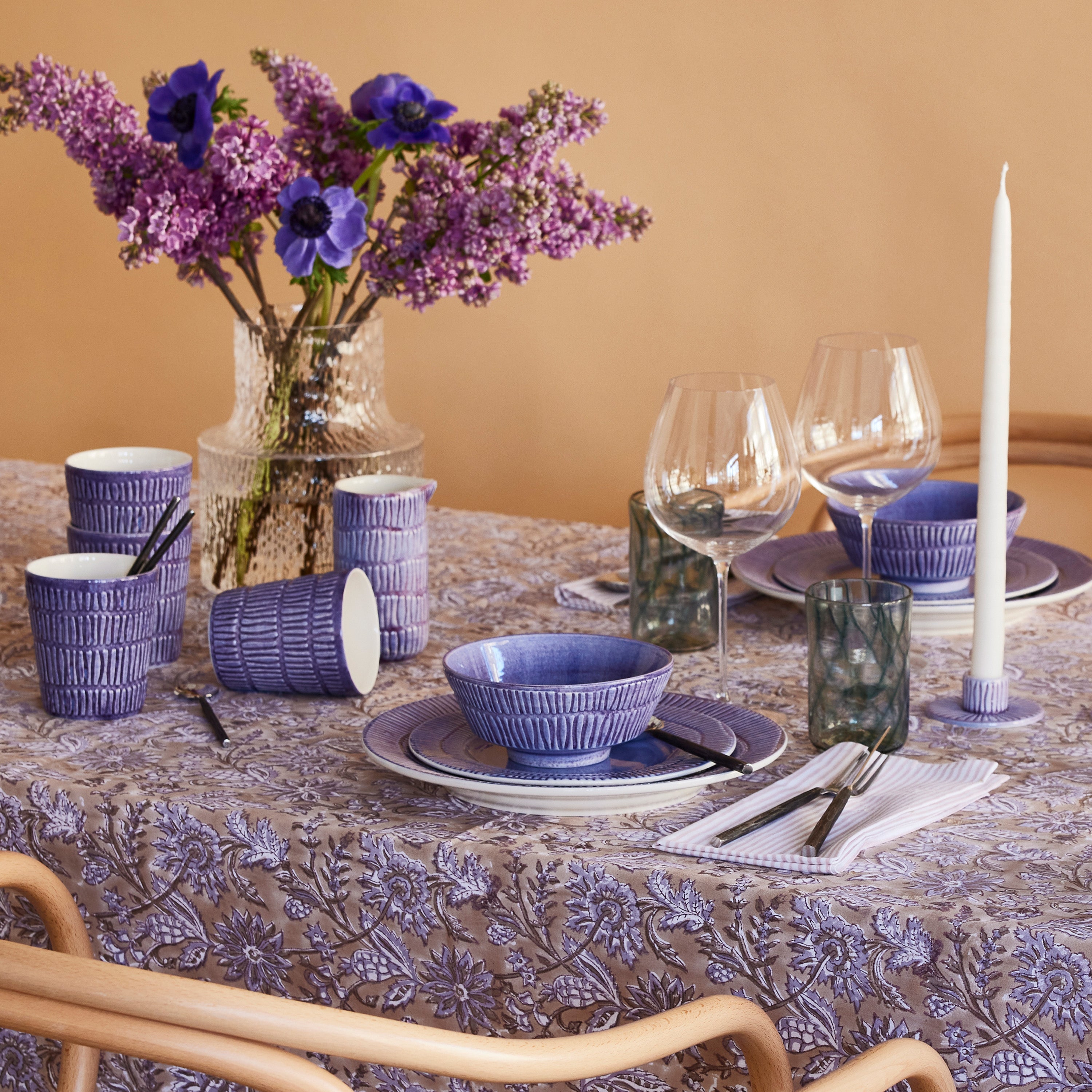 Dining table set with purple and white tableware, flowers, and candles on a patterned tablecloth.