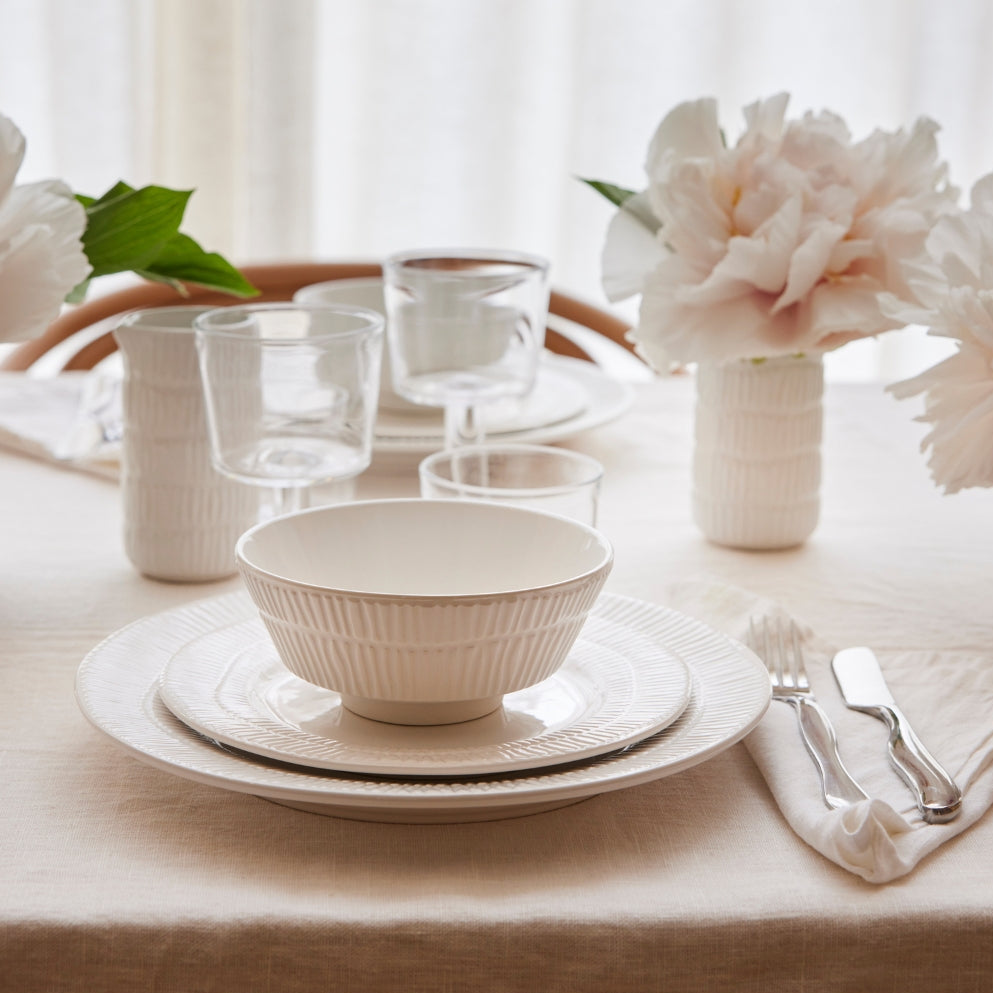 Dining table setting with white ceramic dishes, glasses, and silverware on a beige tablecloth.