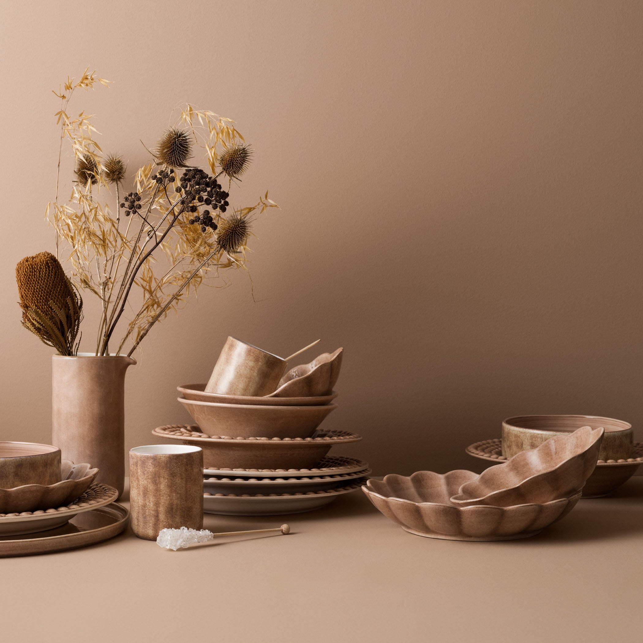 Set of brown ceramic dishes and a vase with dried plants on a beige surface.
