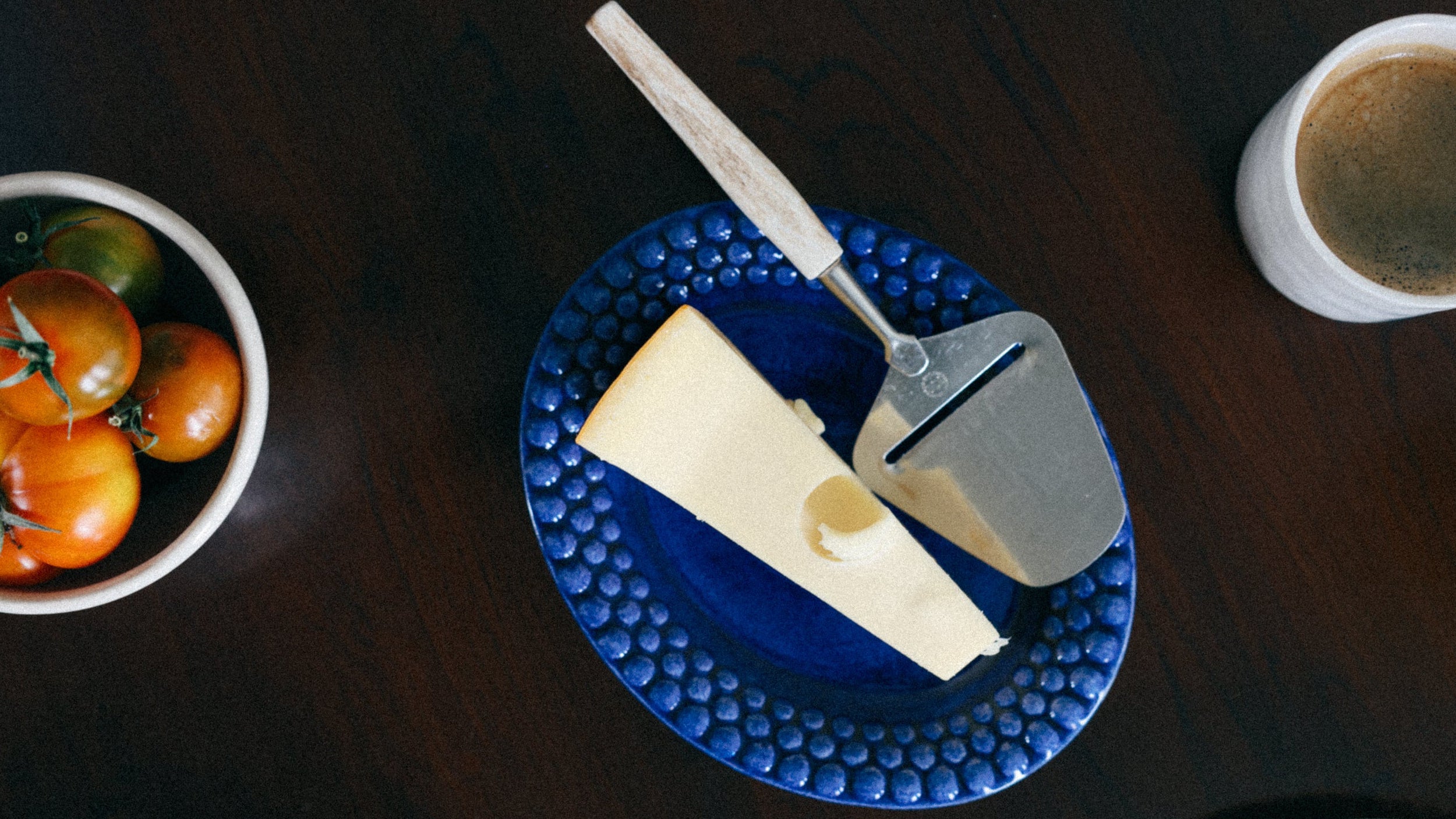 Blue plate with a block of cheese, cheese knife, and bowl of tomatoes on a wooden table.